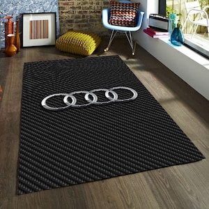 May include: Black rectangular rug with a textured pattern and a silver Audi logo. The rug is placed on a wooden floor in a living room setting. The Audi logo is made of silver metal.