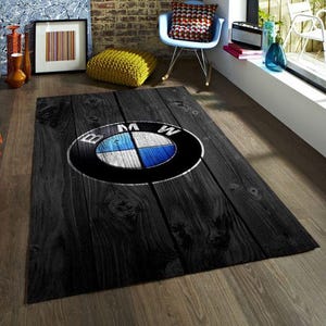 Op de afbeelding: Een rechthoekig zwart tapijt met een houtnerfpatroon. Het tapijt heeft het BMW-logo in het midden, met een blauw-wit embleem in een zwarte cirkel. Het tapijt ligt op een houten vloer.