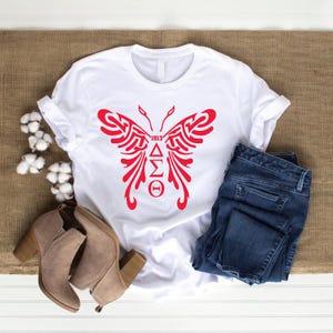 May include: A white t-shirt featuring a red butterfly design with the text "1913" and Greek letters. The shirt is paired with blue jeans and beige ankle boots. The image is set on a neutral background.