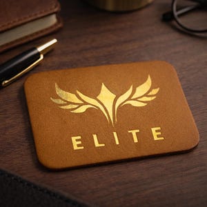 Op de afbeelding: Een bruine leren visitekaarthouder met het woord "ELITE" in gouden letters. De kaarthouder heeft een gouden embleemontwerp. Een zwarte pen, notitieboek en horloge zijn ook zichtbaar.