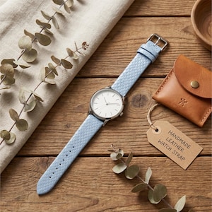 Peut inclure: Une montre-bracelet avec un boîtier rond argenté, un cadran blanc et un bracelet en cuir texturé bleu clair. Une étiquette en carton beige attachée à la montre indique HANDMADE LEATHER WATCH STRAP. Une petite pochette en cuir beige est incluse.