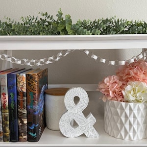 May include: A white bookshelf with books, decorative flowers, and a paper chain. The books are from the Harry Potter series. A white ampersand decorates the shelf. Pink and white flowers are in a white geometric vase.