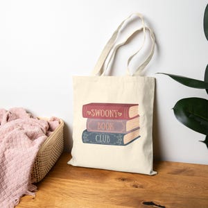 Op de afbeelding: Een crèmekleurige canvas draagtas met een afbeelding van drie gestapelde boeken. Het bovenste boek is rood met de tekst "SWOONY", het middelste is lichtpaars met "BOOK" en het onderste is blauw met "CLUB". Elk boek heeft decoratieve krullen.