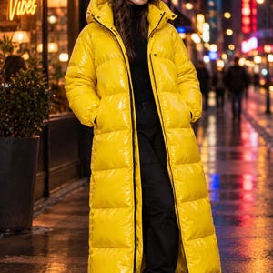 Puede incluir: Un abrigo largo acolchado de color amarillo brillante con un acabado lustroso y barnizado. Esta chaqueta con capucha presenta una cremallera frontal completa, cuello alto y bolsillos laterales profundos. El abrigo llega hasta la parte baja de la pantorrilla.