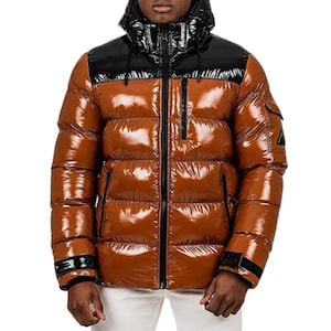 May include: A shiny, brown puffer jacket with a black hood and shoulder panel. The jacket has a zip-up front, multiple pockets, and black accents. The jacket is designed for cold weather.