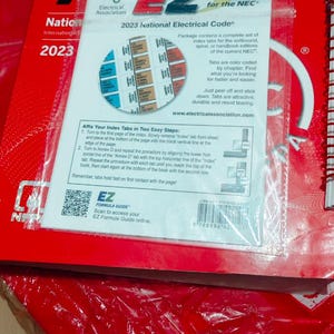 May include: A red book with the text "NFPA 70" and "2023 National Electrical Code" is shown. A package of "EZ Tabs for the NEC" is on top. The tabs are color-coded and designed for the softbound edition.
