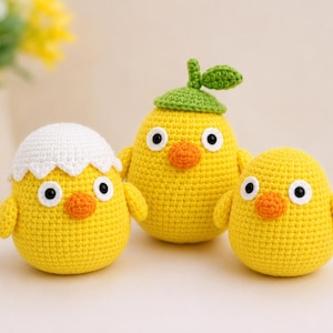 Op de afbeelding: Drie gele gehaakte knuffels met ronde lichamen, oranje snavels en zwart-witte ogen. Eén figuur draagt een wit gehaakt eierschaalmutsje, één een groen gehaakt blaadjesmutsje en de derde is een eenvoudig geel kuikentje.