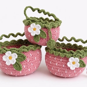 Op de afbeelding: Drie ronde gehaakte manden met een roze aardbeienpatroon en witte verticale streepjes. Elke mand heeft een groene geschulpte rand, een groen golvend handvat en een witte bloemapplicatie met een geel hartje en groene blaadjes.