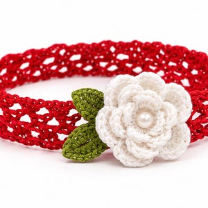 Könnte beinhalten: Ein rotes Häkelstirnband mit durchbrochenem Muster, verziert mit einer weißen Häkelblume mit einer zentralen Perle und zwei kleinen grünen Häkelblättern, die an der Seite des Bandes befestigt sind.