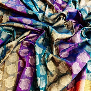 May include: A vibrant, patterned textile with a rich color palette of teal, purple, gold, and yellow. The fabric features a repeating paisley design. The cloth is draped, creating folds and shadows that enhance the texture and visual interest.