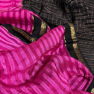 May include: A vibrant textile featuring a striking contrast of colors and patterns. The image showcases a section of fabric with bold pink stripes, juxtaposed against a black background with a gold grid design. The edges are trimmed with black and gold detailing.