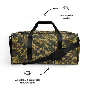 Könnte beinhalten: Eine Camouflage-Duffel-Tasche mit einem sich wiederholenden Smiley-Gesicht-Design in Gelb und Braun. Die Tasche hat schwarze Griffe und einen verstellbaren, abnehmbaren Schultergurt. Der Text lautet "Dual padded handles" und "Adjustable & removable shoulder strap".