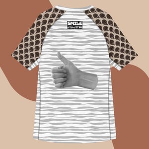 May include: A white and gray rash guard with a thumbs-up graphic. The sleeves have a brown pattern. The shirt has the text "SMILE JIU-JITSU".