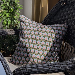 May include: A decorative throw pillow with a blue, brown, and pink floral pattern rests on a black wicker chair. The pillow's design features a repeating pattern of stylized flowers. A small green plant is visible to the left of the chair.