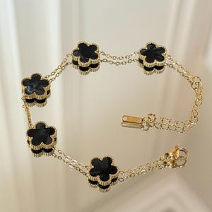 Puede incluir: Una pulsera dorada con seis abalorios negros en forma de trébol. Los abalorios están conectados por delicadas cadenas doradas. La pulsera tiene un cierre y una cadena ajustable. La pulsera se muestra sobre una superficie reflectante.