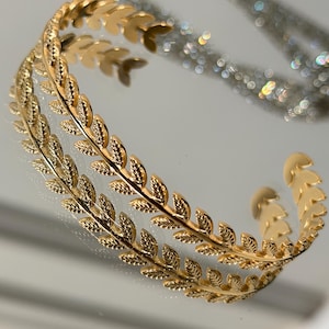 May include: A gold-coloured cuff bracelet with a leaf design. The bracelet is open at the ends and features a repeating pattern of leaves. The bracelet is resting on a reflective surface, with a blurred, sparkly background.