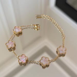 May include: A gold-toned chain bracelet featuring five evenly spaced flower-shaped charms. Each charm contains a smooth, iridescent, pearlescent inlay. The bracelet includes a delicate link chain and a small lobster claw clasp closure.