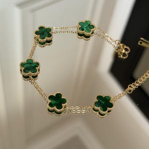 May include: Gold bracelet with six green clover-shaped charms. The charms are linked by a fine chain. The bracelet is shown on a reflective surface, emphasizing its polished appearance. A timeless piece of jewellery.
