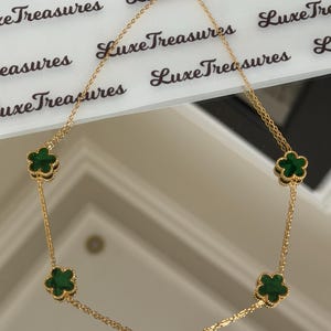 May include: A gold-colored necklace featuring five clover-shaped pendants with a dark green center. The necklace is displayed on a white surface with the text "Luxe Treasures" printed repeatedly.