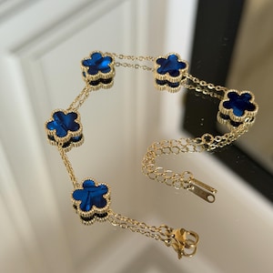 Op de afbeelding: Een goudkleurige armband met vijf klavervormige bedels met een diepblauwe kern. De bedels zijn verbonden door een fijne ketting en de armband heeft een sluiting.