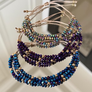 Puede incluir: Tres pulseras de cuentas en tonos azul, morado y verde iridiscente. Cada pulsera presenta múltiples hilos de pequeñas cuentas facetadas y detalles dorados. Las pulseras tienen cordones beige ajustables.