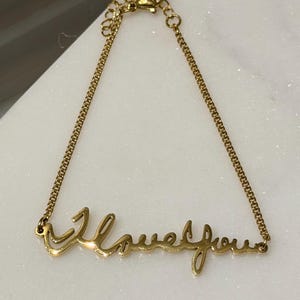 May include: A gold-colored bracelet with the words "I love you" in a cursive font. The bracelet is made of a delicate chain and has an adjustable clasp. The jewelry is displayed on a white surface.