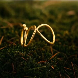 May include: A gold-colored ring with an abstract design, featuring two small spheres and a looping shape. The ring is set against a backdrop of green moss, creating a natural contrast. The ring's design is modern and minimalist.