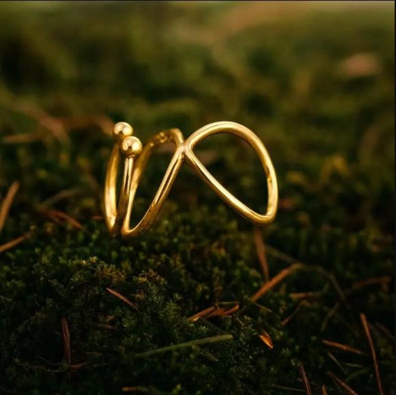 May include: A gold-colored ring with an abstract design, featuring two small spheres and a looping shape. The ring is set against a backdrop of green moss, creating a natural contrast. The ring's design is modern and minimalist.