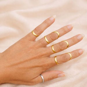 May include: A hand displaying a collection of gold-colored rings. The rings are of varying designs, with several featuring a curved, open-ended shape. One silver ring is also visible. The background is a soft, neutral color.