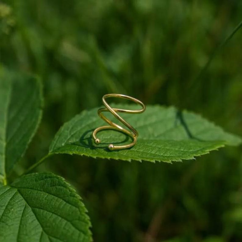 May include: A gold-colored, spiral-shaped ring with a minimalist design. The ring is resting on a green leaf, with a blurred green background. The ring's design features a continuous, looping form.
