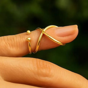 May include: A gold-colored finger ring with an X-shaped design and two small ball accents. The ring is worn on a finger, with a blurred green background.