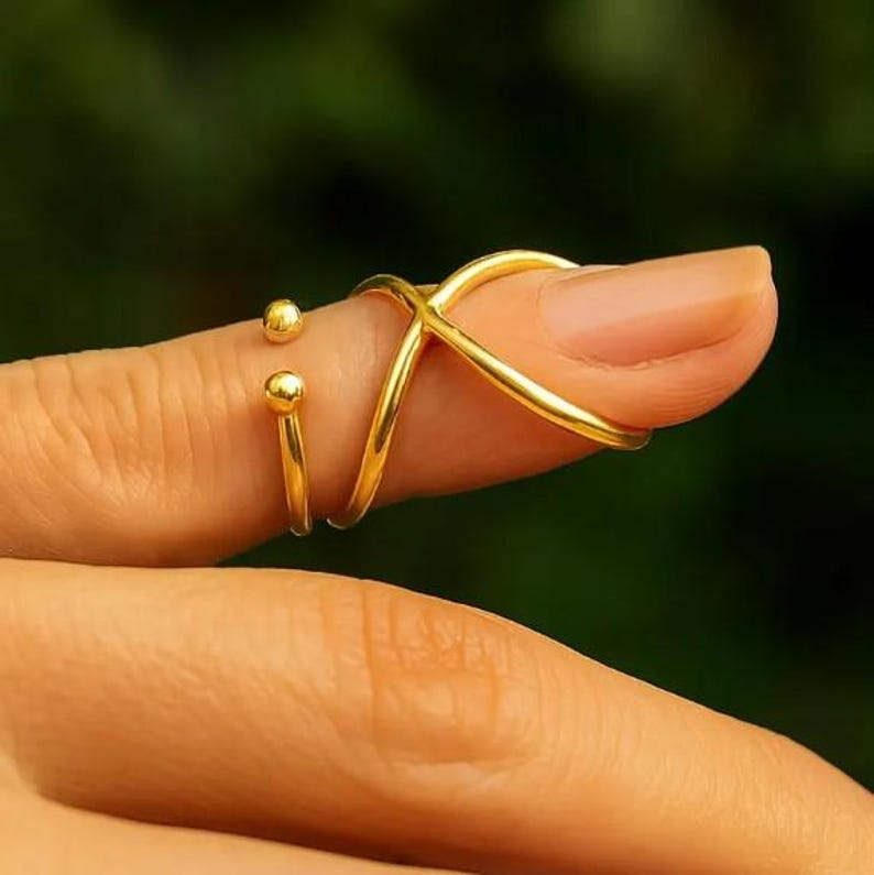 May include: A gold-colored finger ring with an X-shaped design and two small ball accents. The ring is worn on a finger, with a blurred green background.