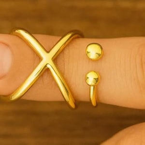 May include: A gold-colored ring set. The set includes an X-shaped ring and two separate rings with a small ball design. The rings are worn on a finger.