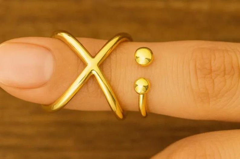 May include: A gold-colored ring set. The set includes an X-shaped ring and two separate rings with a small ball design. The rings are worn on a finger.