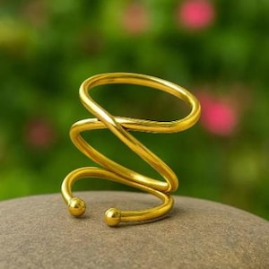 May include: A gold-colored, spiral-shaped ring with a smooth, polished finish. The ring is resting on a gray stone, with a blurred green and pink background. The design features a continuous, looping form.