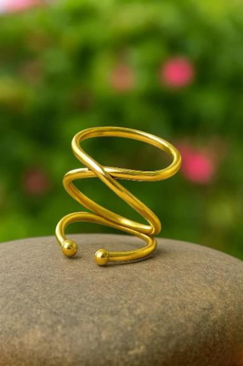 May include: A gold-colored, spiral-shaped ring with a smooth, polished finish. The ring is resting on a gray stone, with a blurred green and pink background. The design features a continuous, looping form.