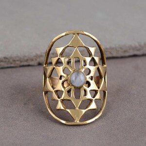 May include: A gold-coloured ring with an intricate geometric design. The ring features an oval shape with a central, light-coloured stone. The design includes triangles and other geometric shapes, creating a detailed and ornate appearance.