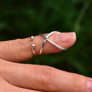May include: A silver-colored finger ring with an X-shaped design and two small, round accents. The ring is worn on a finger, with a blurred green background.