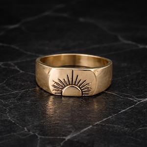 May include: A gold-colored ring with a rectangular face featuring a sunburst design. The ring has a thick band and a slightly textured surface. The sunburst design is etched into the face of the ring.