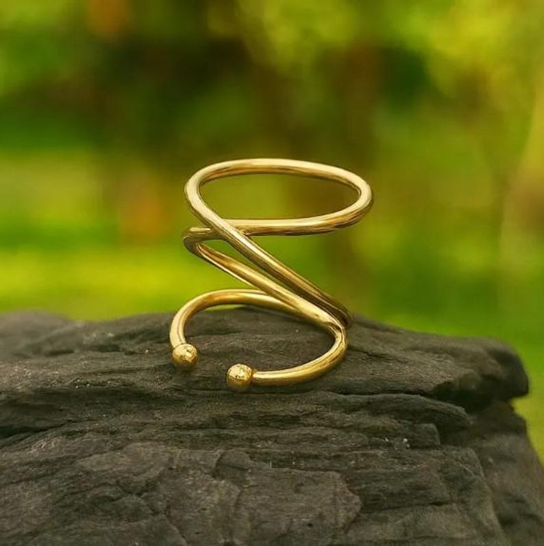 May include: A gold-colored ear cuff with a spiral design. The ear cuff has a curved shape with two small ball-shaped ends. The jewelry is displayed on a dark gray rock with a blurred green background.