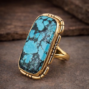 May include: A gold-toned ring featuring a large, rectangular turquoise stone. The turquoise has a striking blue-green color with dark matrix patterns. The ring's setting has a detailed, textured design.