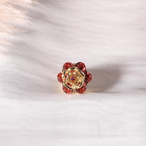 May include: A gold-toned ring with a rose design. The ring features a central cluster of small, pink crystals surrounded by larger, red crystals. The petals are detailed with a gold finish, creating a floral jewelry piece.