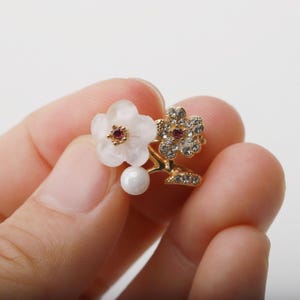以下が含まれることがあります： 2つの花のモチーフが特徴のゴールドトーンのフローラルリング。1つの花は白で、赤い中心と白いパールが付いています。もう1つの花は、透明なクリスタルと赤い中心がちりばめられています。リングは人が持っています。