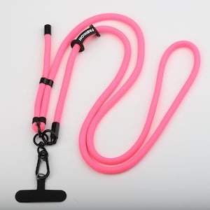 Könnte beinhalten: Leuchtend pinkfarbenes Handy-Lanyard mit schwarzem Karabiner und Stecker. Auf einem schwarzen Band steht das Wort "FASHION". Das Band besteht aus einem dicken, gewebten Material.