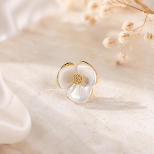 Pode incluir: Um brinco de pino dourado em forma de flor de três pétalas. As pétalas são feitas de material de concha branca iridescente e o centro apresenta um pequeno detalhe de estame dourado texturizado com um acabamento metálico polido.