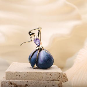 Puede incluir: Broche de bailarina dorada con una gema púrpura y una falda azul y morada. El broche está colocado sobre un bloque de piedra clara sobre un fondo suave y neutro. La bailarina está en una pose elegante.
