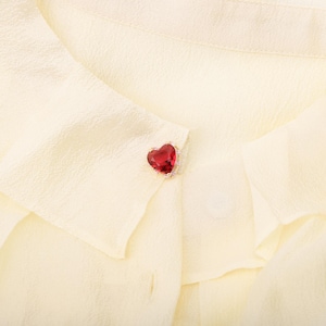 May include: Close-up of a cream-colored blouse with a heart-shaped red jewel brooch. The brooch is set in a gold-colored metal and surrounded by small, clear stones. The blouse has a textured fabric.