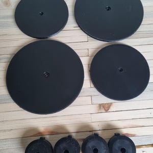 May include: A set of black, circular components, comprising four large discs and four smaller, cylindrical parts. The discs feature a central aperture. The items are displayed on a light-coloured wooden surface.
