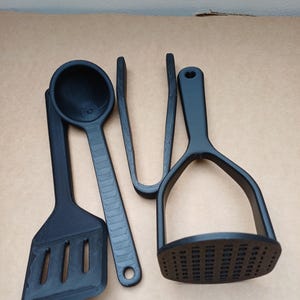 May include: A collection of four black kitchen tools: a slotted spatula, a ladle, tongs, and a potato masher. The utensils are made of a matte black material and are arranged on a neutral-toned surface.