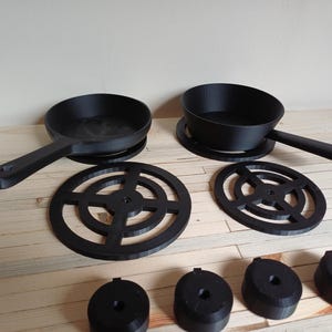 May include: A black miniature cookware set, comprising two frying pans with handles, two circular trivets, and four small black components. The pans and trivets are round, and the set is arranged on a light wood surface.